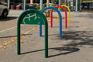 Ein Fahrradbügel SALM mit Schild in Moosgrün montiert als Reihenanlage an einer Straße mit anderen Anlehnbügeln SALM ohne Schild in Enzianblau, Verkehrsrot und einer Sonderfarbe Gelb