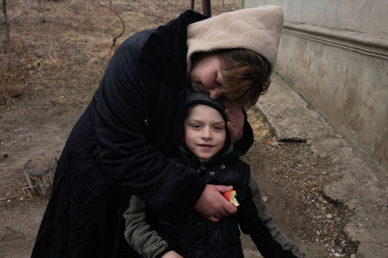 Auf einer zerstörten ukrainischen Straße umarmt eine Frau mit tragenden beigen Pullover und einer großen, dicken schwarzen Jacke einen kleinen Jungen mit grauer Jacke und einer grauen Mütze.
