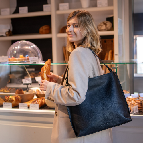 FEYNSINN Leder schwarz JUNA Outdoor Frau steht im Kaffee und haelt ein croissant in der Hand