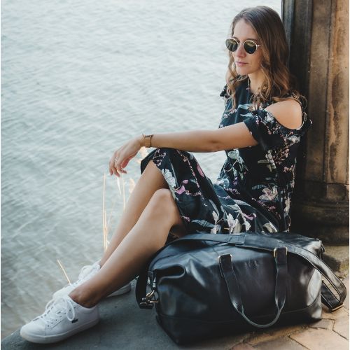 Sid & Vain Reisetasche aus Leder in schwarz Outdoor Frau sitzt am Wasser und traegt ein sommerliches dunkles Kleid