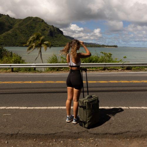 SONS OF ALOHA 2in1 Rucksack und Trolley Outdoor am Meer Frau steht am Straßenrand und schaut aufs Meer