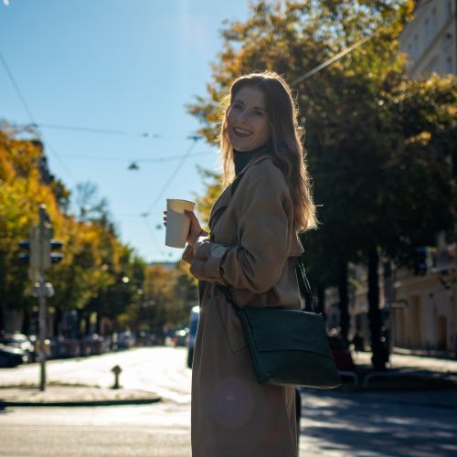 FEYNSINN Umhaengetasche LIVA leder in dunkegruen Frau steht draußen und traegt einen trenchcoat und haelt einen kaffee in der hand