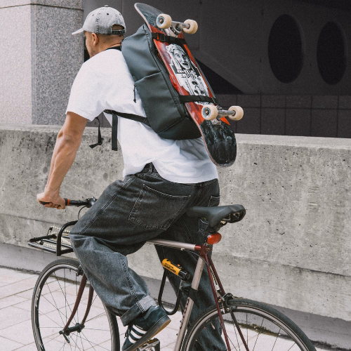SONS OF ALOHA Uni Rucksack Herren AHI in schwarz Outdoor Mann faehrt Fahrrad und traegt ein weißes oversized T-Shirt mit einer weiten grauen Jeans
