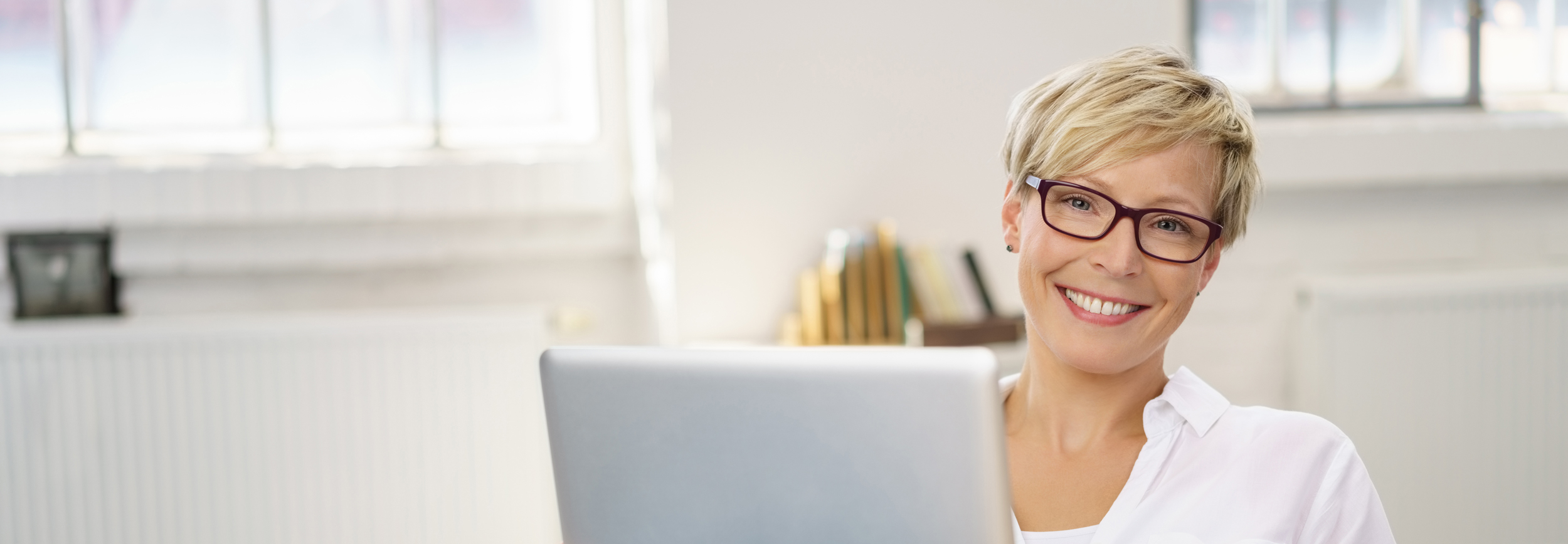 Porträt einer lächelnden Frau mit Brille, die an einem Laptop arbeitet und Freundlichkeit und Servicebereitschaft vermittelt.