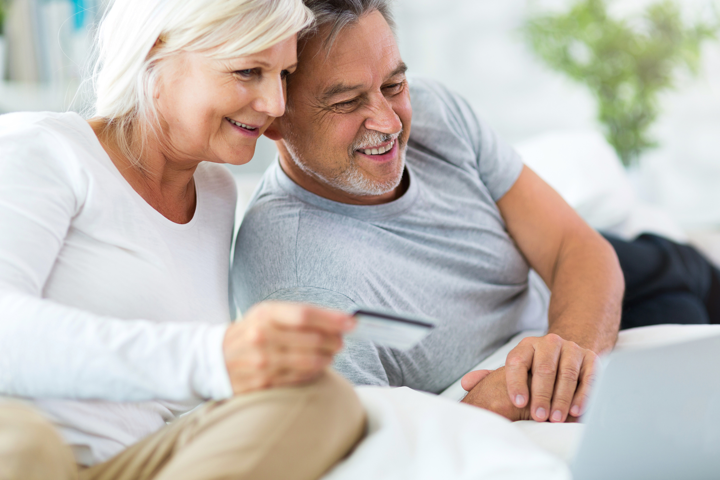 Ein älteres Paar lacht, während es gemeinsam auf einem Laptop online einkauft. Die Frau hält eine Kreditkarte zur Bezahlung.