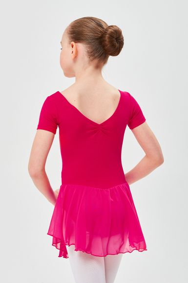 Ein junges Mädchen in einem rosa Ballettkleid und weißen Strumpfhosen steht mit abgewandtem Gesicht und den Händen hinter dem Rücken da.