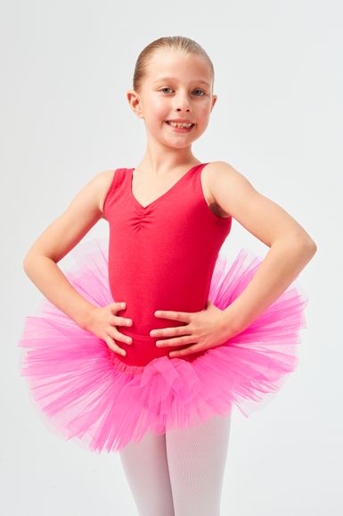 Ein junges Mädchen in einem rosa Tutu und einem roten Ballettbody steht mit den Händen in den Hüften und lächelt in die Kamera.