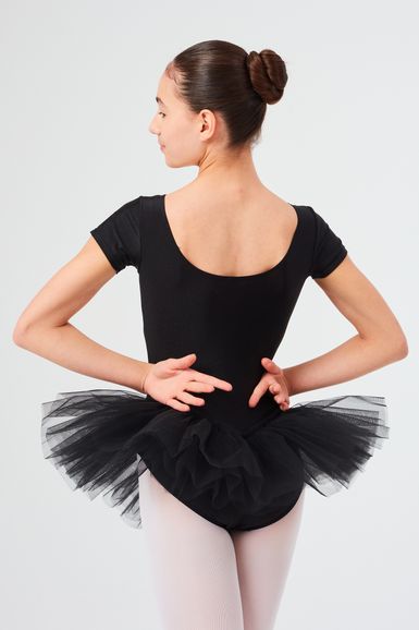 Eine Balletttänzerin in einem schwarzen Ballettbody und Tutu steht mit den Händen in den Hüften und blickt von der Kamera weg.