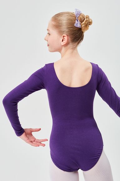 Ein junges Mädchen in einem lila Ballettbody und einer Schleife steht in einer Ballettpose und blickt von der Kamera weg.