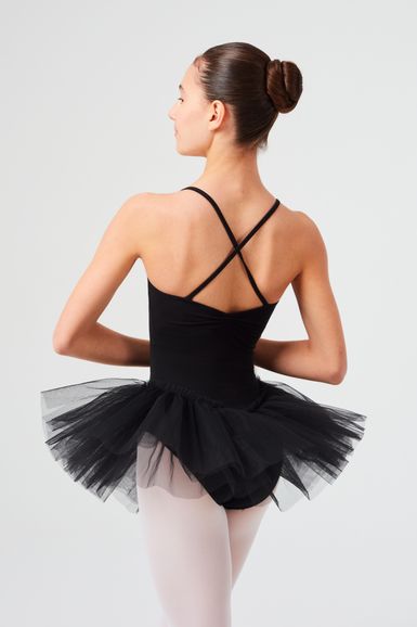 Eine Balletttänzerin in einem schwarzen Ballettbody und Tutu steht mit in die Hüften gestemmten Armen und blickt von der Kamera weg.