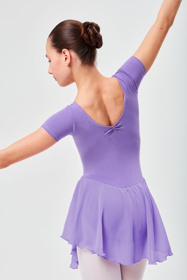 Eine junge Balletttänzerin in einem lavendelfarbenen Ballett Trikot und Rock posiert mit ausgestreckten Armen und blickt von der Kamera weg.
