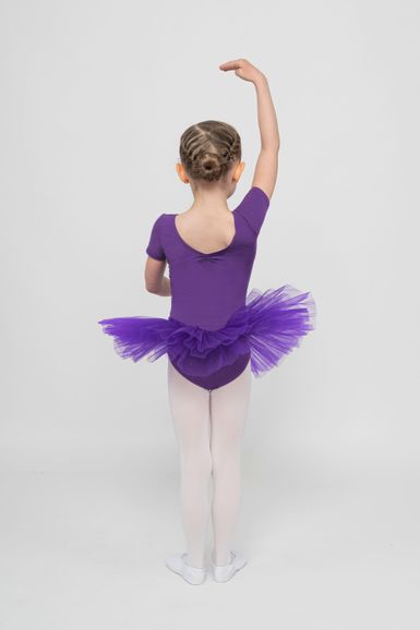 Ein kleines Kind in einem lila Ballett-Outfit und weißen Strumpfhosen posiert mit einem erhobenen Arm und blickt von der Kamera weg.