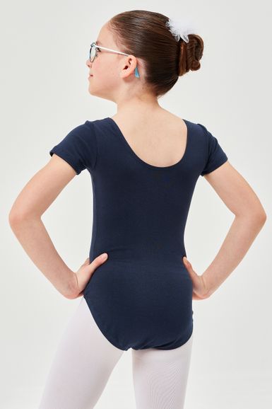 Ein Mädchen in einem marineblauen Ballett Trikot und weißen Strumpfhosen steht mit den Händen in den Hüften und blickt von der Kamera weg.