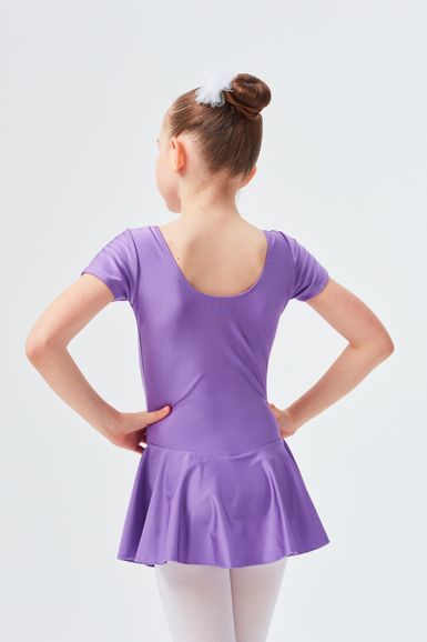 Ein junges Mädchen in einem lila Ballett Trikot und weißen Strumpfhosen steht mit den Händen in den Hüften und blickt von der Kamera weg.