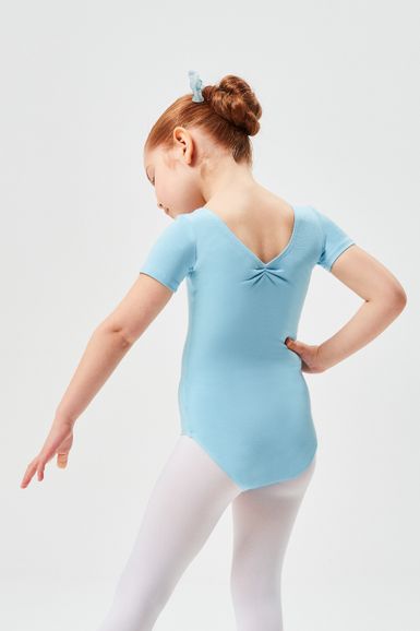 Ein junges Mädchen in einem hellblauen Ballett Trikot und weißen Strumpfhosen steht mit einer Hand auf der Hüfte und blickt von der Kamera weg.