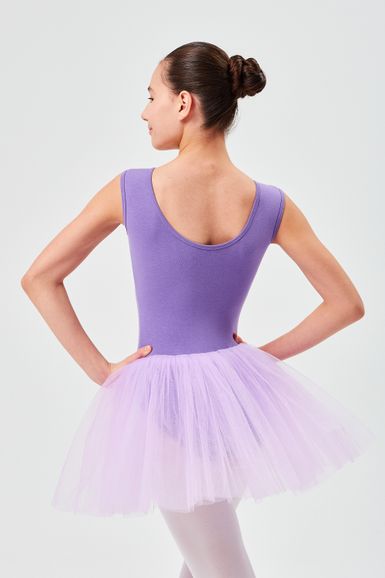 Eine Balletttänzerin in einem lila Ballett Trikot und Tutu steht mit den Händen in den Hüften und blickt von der Kamera weg.