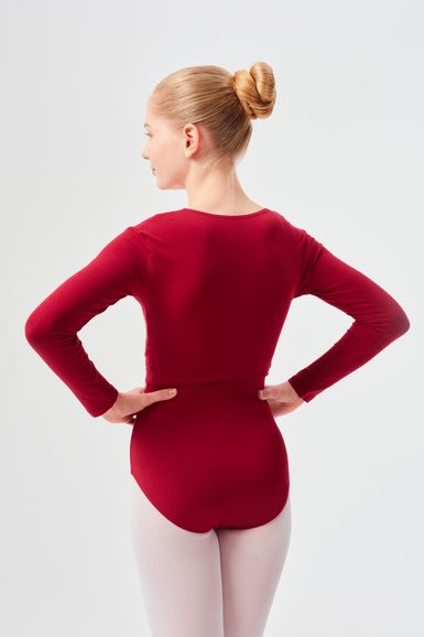 Eine junge Frau in einem roten Ballett Trikot und weißen Strumpfhosen steht mit abgewandtem Gesicht und den Händen in den Hüften.