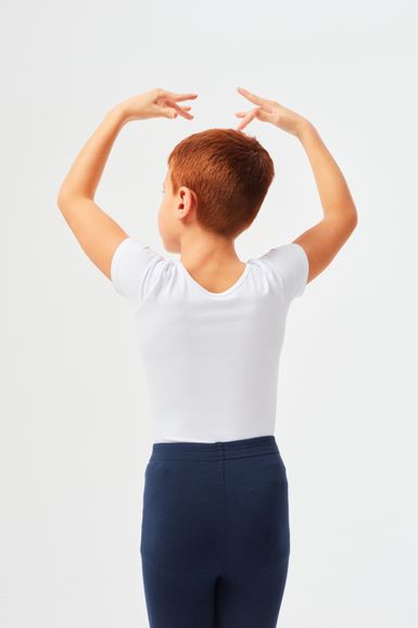 Ein Kind mit kurzen roten Haaren in einem weißen Oberteil und einer dunkelblauen Hose steht mit abgewandtem Gesicht und erhobenen Armen in einer Ballettpose da.