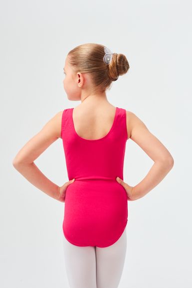 Mädchen in einem rosa Ballett Trikot und weißen Strumpfhosen steht mit den Händen in den Hüften und blickt von der Kamera weg.