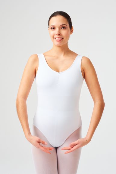 Eine Frau in einem weißen Trikot und Strumpfhosen steht in der ersten Ballettposition mit nach vorne gebeugten Armen und lächelt in die Kamera.