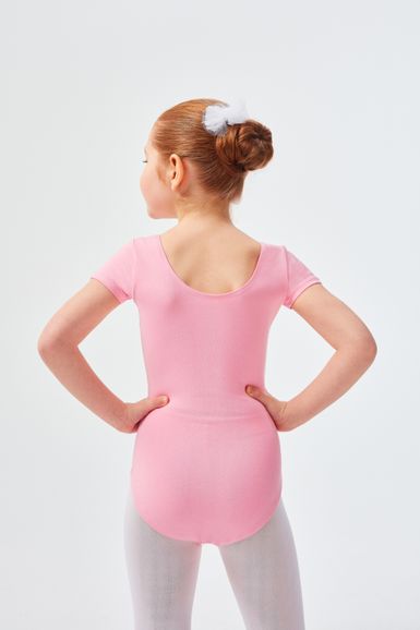 Ein junges Mädchen in einem rosa Ballettbody und weißen Strumpfhosen steht mit den Händen in den Hüften und blickt von der Kamera weg.