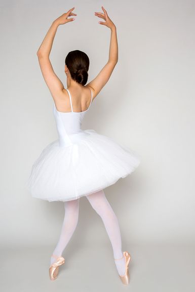 Eine Balletttänzerin in einem weißen Tutu und Spitzenschuhen steht mit erhobenen Armen auf Spitze und blickt von der Kamera weg.