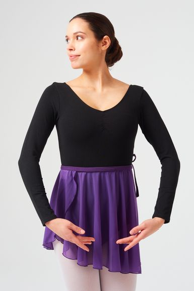 Eine Frau in einem schwarzen Trikot und einem lila Rock steht in einer Ballettpose mit vor ihr verschränkten Armen.