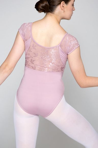 Eine Balletttänzerin in einem hellrosa Trikot mit Spitzenrücken und weißen Strumpfhosen posiert mit leicht angewinkelten Armen und blickt von der Kamera weg.