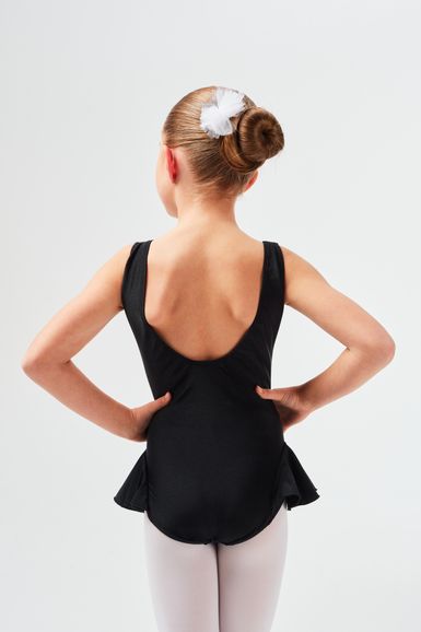 Ein junges Mädchen in einem schwarzen Ballettbody und weißen Strumpfhosen steht mit den Händen in den Hüften und blickt von der Kamera weg.