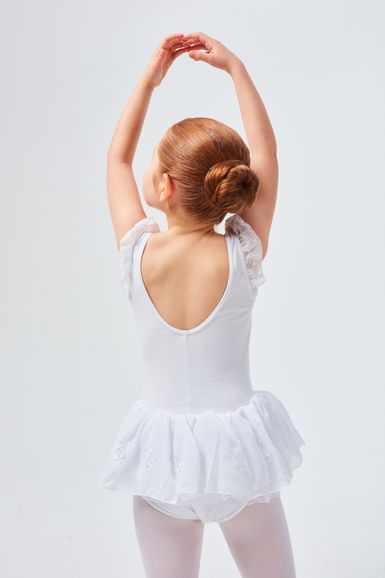 Ein junges Mädchen in einem weißen Ballett-Outfit steht mit über dem Kopf erhobenen Armen da und blickt von der Kamera weg.