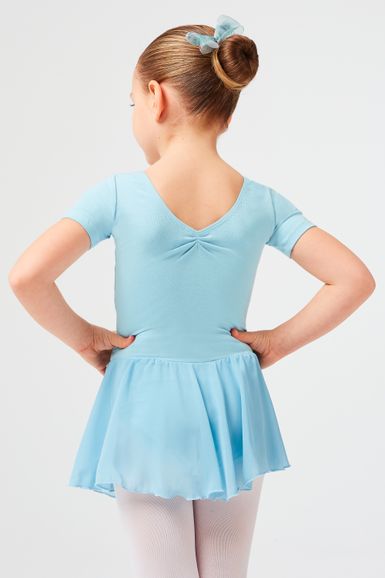 Ein junges Mädchen in einem hellblauen Ballett-Outfit steht mit den Händen in den Hüften und blickt von der Kamera weg.