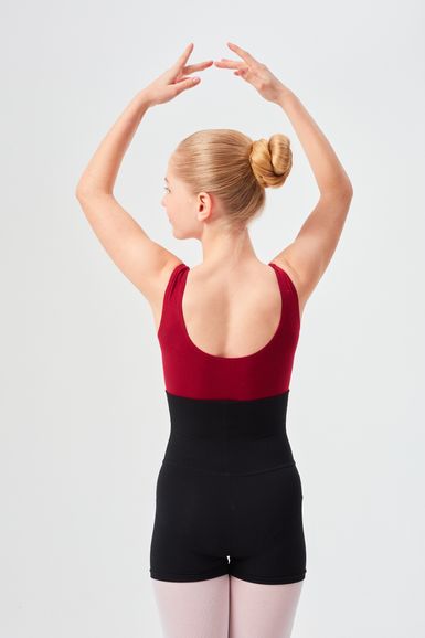 Junge Balletttänzerin in einem roten Trikot und schwarzen Shorts posiert mit erhobenen Armen und dem Rücken zur Kamera.