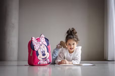 Kind liegt am Boden und zeichnet, daneben ein rosa Minnie‑Rucksack mit großem Motiv und Herzschleife.