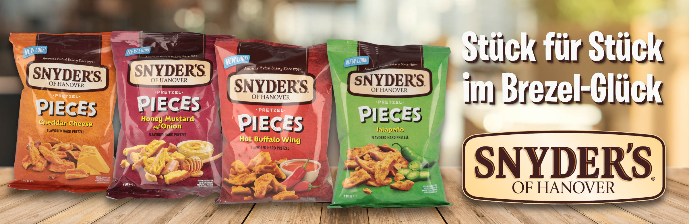 Das Bild zeigt vier verschiedene Sorten von „Snyder's of Hanover Pretzel Pieces“ aufgereiht auf einer Holzoberfläche. Von links nach rechts sind die Geschmacksrichtungen Cheddar Cheese, Honey Mustard & Onion, Hot Buffalo Wing und Jalapeño zu sehen. Rechts neben den Tüten steht der Slogan „Stück für Stück im Brezel-Glück“ über dem Logo von Snyder's of Hanover. Der Hintergrund ist unscharf und warm beleuchtet.