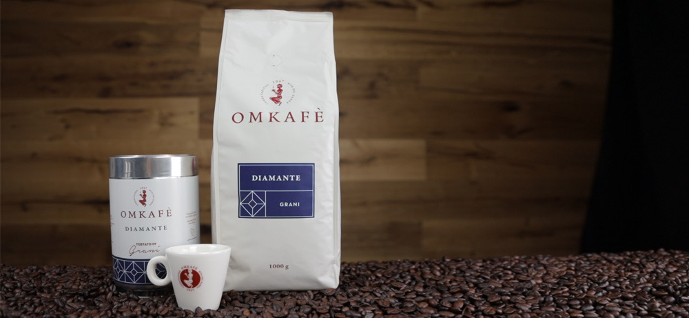 Omkafe Diamante 1000g Bohnenpackung mit Diamante-Vorratsdose und Omkafe-Espressotasse vor dunklem Holzhintergrund auf Kaffeebohnen