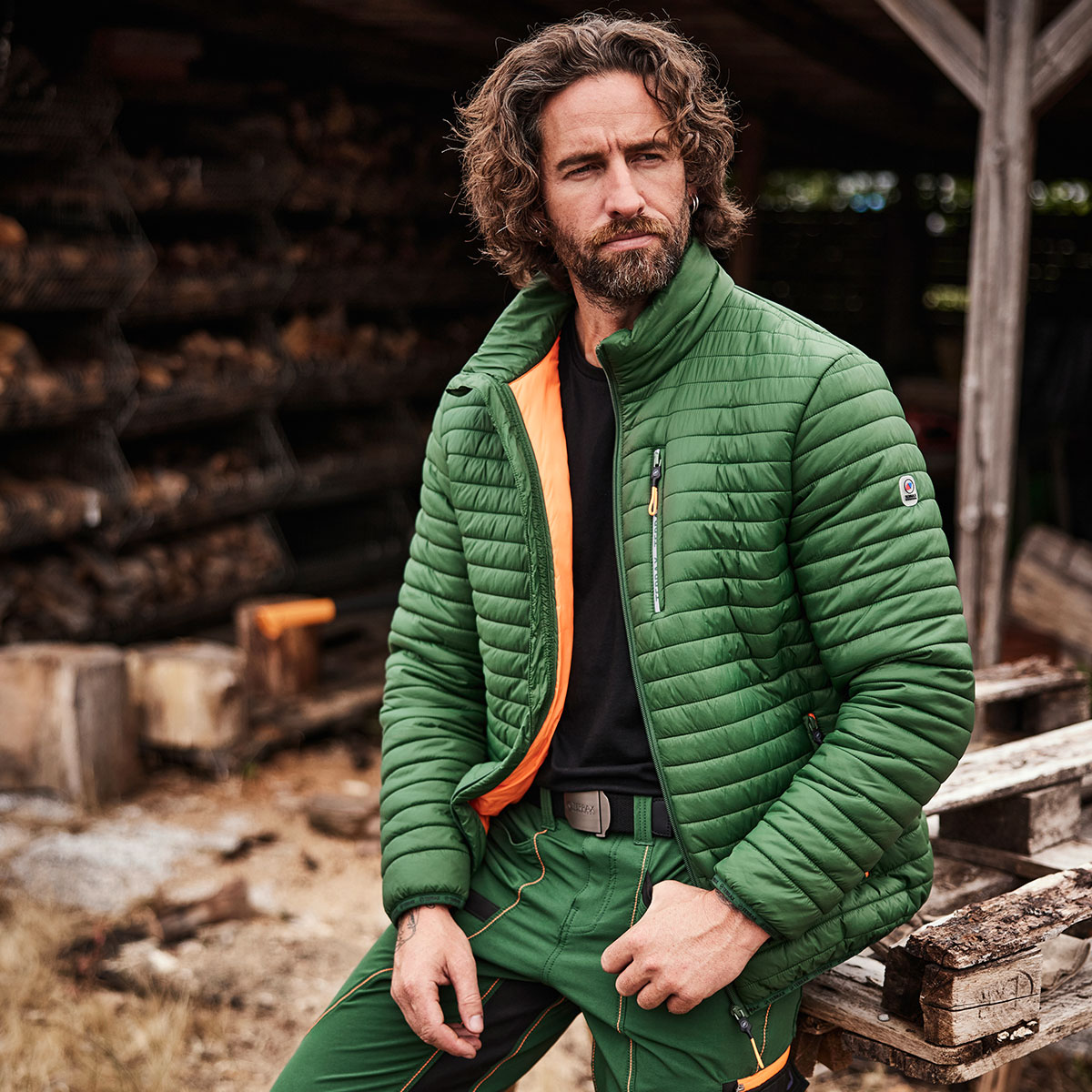 Mann steht mit einer Steppjacke von Terrax Workwear vor einem Arbeitsbereich für Brennholz
