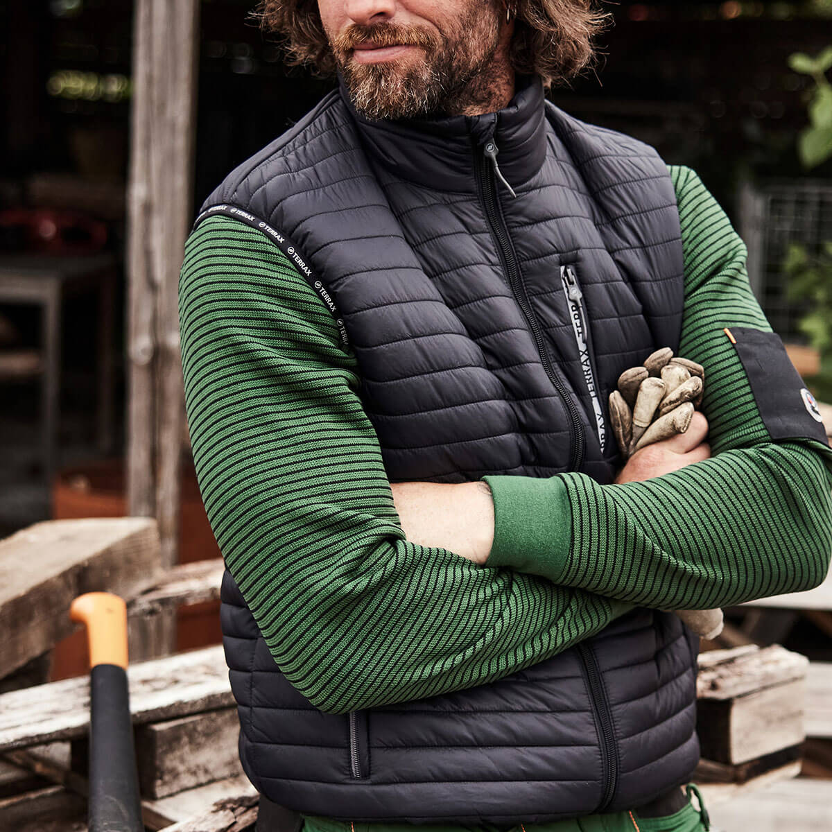 Mann mit verschränkten Armen trägt eine Weste von Terrax Workwear vor einem Arbeitsbereich für Brennholz