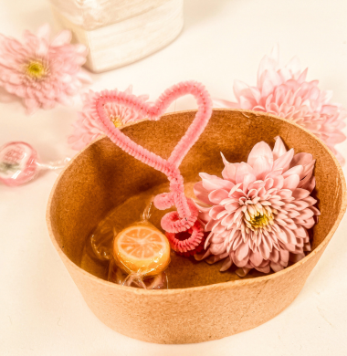 Eine geöffnete Pappschachtel, im Inneren ein Herz aus Chenille, eine Blume und ein Bombon.