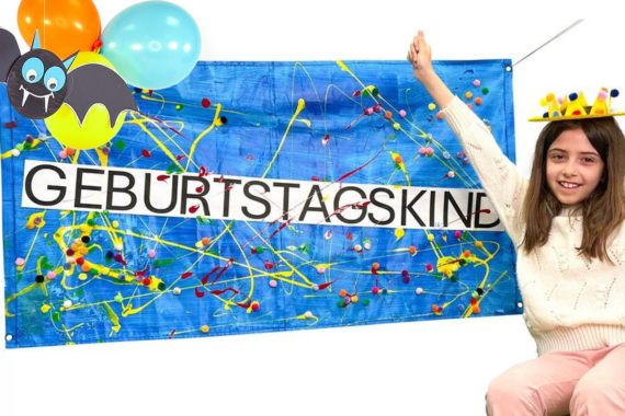 Ein großer Geburtstagsbanner, der mit Farben, Pompons und Luftballons sowie einer Fledermaus aus Tonpapier geschmückt ist. Auf der rechten Seite sitzt ein Mädchen, das freudig die Arme gehoben hat und in die Kamera lächelt. Auf dem Kopf trägt sie eine Geburtstagskrone aus einem Pappteller gebastelt. Auf dem Bannersteht in Großbuchstaben "Geburtstagskind".