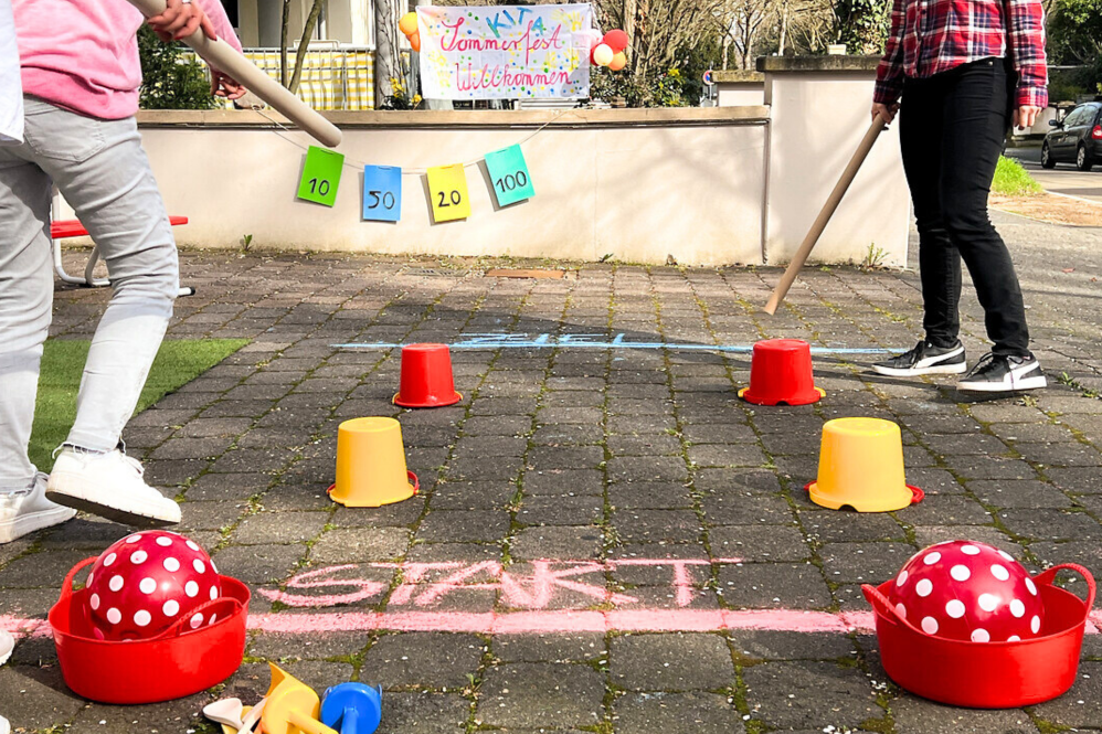 Outdoor-Aufnahme: Auf dem Boden wurde mit Kreide eine Start- und eine Ziellinie gemalt. Mit Eimern wurde ein Parkour für zwei Teilnehmer gebaut. Diese schieben mit einem langen Pappstab jeweils ihren Ball durch den Parkour.