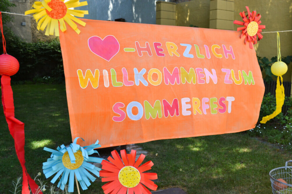 Ein großer orangenfarbiger Banner mit einer bunten Aufschrift: "Herzlich willkommen zum Sommerfest".