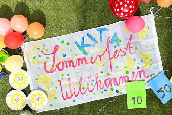 Ein großer Banner mit der Aufschrift: "Kita Sommerfest Willkommen", daneben liegen Luftballons, beschmückte Pappteller- und Moosgummi-Spiele und ein Ball.