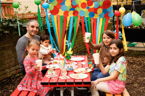 Vier Kinder und ein Erwachsener sitzen an einem roten Tisch im Freien und lachen in die Kamera. Die Szene ist sommerlich-festlich. Der Hintergrund ist geschmückt mit Luftballons und einer Papiergirlande.