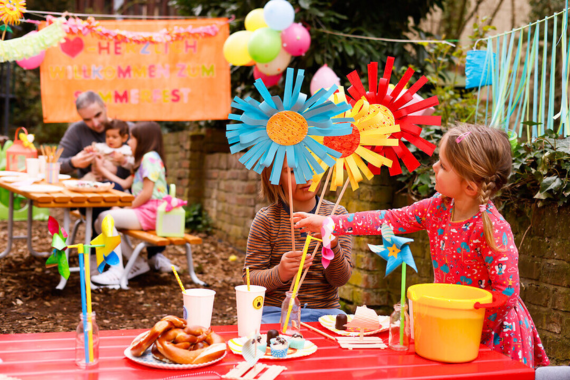 Outdoor-Szene: Sommerfest. Zwei Kinder spielen im Vordergrund mit gebastelten Blumen, vor ihnnen ein geschmückter Tisch. Im Hintergrund mehr Deko  und ein Mann mit zwei Kindern.