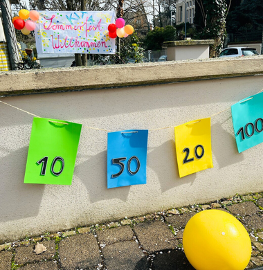 Gebastelte Moodgummi-Zielscheiben hängen an einer Mauer. Ein gelber Ball liegt davor. Im Hintergrund sieht man einen großen Banner mit der Aufschrift "Kita Sommerfest Willkommen".