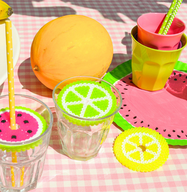 Sommerlich geschmückter Tisch. Im Fokus sind Gläser, die mit Bügelperlen-Platten in Obst-Optik verziert sind. Diese liegen als Deckel auf dem jeweiligen Glas und haben in der Mitte ein Loch, durch den ein Strohalm gesteckt wird.