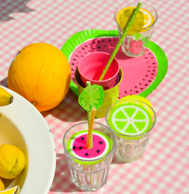 Sommerlich geschmückter Tisch. Im Fokus sind Gläser, die mit Bügelperlen-Platten in Obst-Optik verziert sind. Diese liegen als Deckel auf dem jeweiligen Glas und haben in der Mitte ein Loch, durch den ein Strohalm gesteckt wird. Im Hintergrund sieht man auch einen Pappteller, der angemalt wurde wie eine Wassermelonenscheibe.