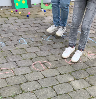 Gemalte Kreidemuster auf dem Boden, die einen zweispurigen Parkour bilden: Jeweils ein Kreis mit einer Linie, die den Kreis halbiert.  Auf der rechten Seite des Bildes sieht man die Füße zweier Personen, die den Parkour durchqueren.