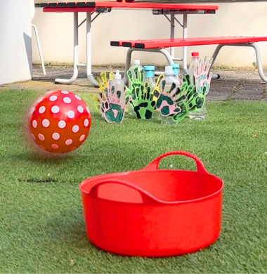 Ein großer Eimer im Vordergrund. Im Hintergrund sieht man wie ein Ball auf selbstgebastelte Kegel zurollt. Die Kegel bestehen aus Plastikflaschen, die mit Händen aus Tonpapier dekoriert sind.
