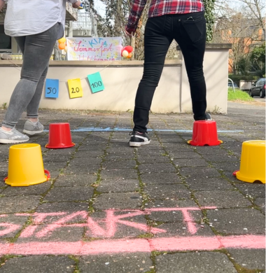 Outdoor-Aufnahme: Auf dem Boden wurde mit Kreide eine Start- und eine Ziellinie gemalt. Mit Eimern wurde ein Parkour für zwei Teilnehmer gebaut. Diese bewegen sich auf die Ziellinie zu.